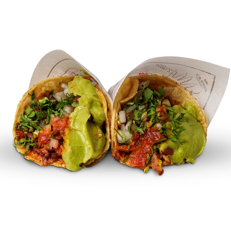 Two adobada tacos in paper wrappers, topped with guacamole, cilantro and salsa